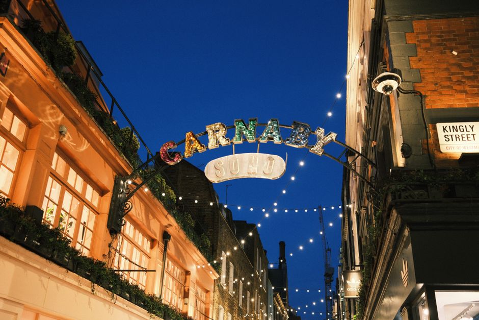 The image shows a busy street scene on Carnaby Street, London, with various commercial buildings on either side, including a MAC cosmetics store visible on the left. Overhead, there are decorative gol
